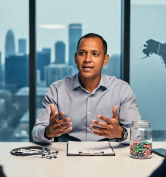 IMG public health physician at a conference table with stethoscope and glass jar of poker chips in the foreground, colleagues blurred, soft daylight, Toronto skyline visible in background.